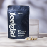 A stand-up pouch of feelgüd Dual Magnesium Capsules stands on a light surface beside a clear glass of water, with a few capsules placed in front. The pouch is dark navy with large white vertical 'feelgüd' branding and white product text.