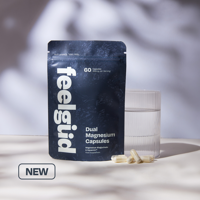 A stand-up pouch of feelgüd Dual Magnesium Capsules stands on a light surface beside a clear glass of water, with a few capsules placed in front. The pouch is dark navy with large white vertical 'feelgüd' branding and white product text.