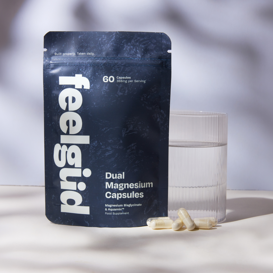 A stand-up pouch of feelgüd Dual Magnesium Capsules stands on a light surface beside a clear glass of water, with a few capsules placed in front. The pouch is dark navy with large white vertical 'feelgüd' branding and white product text.